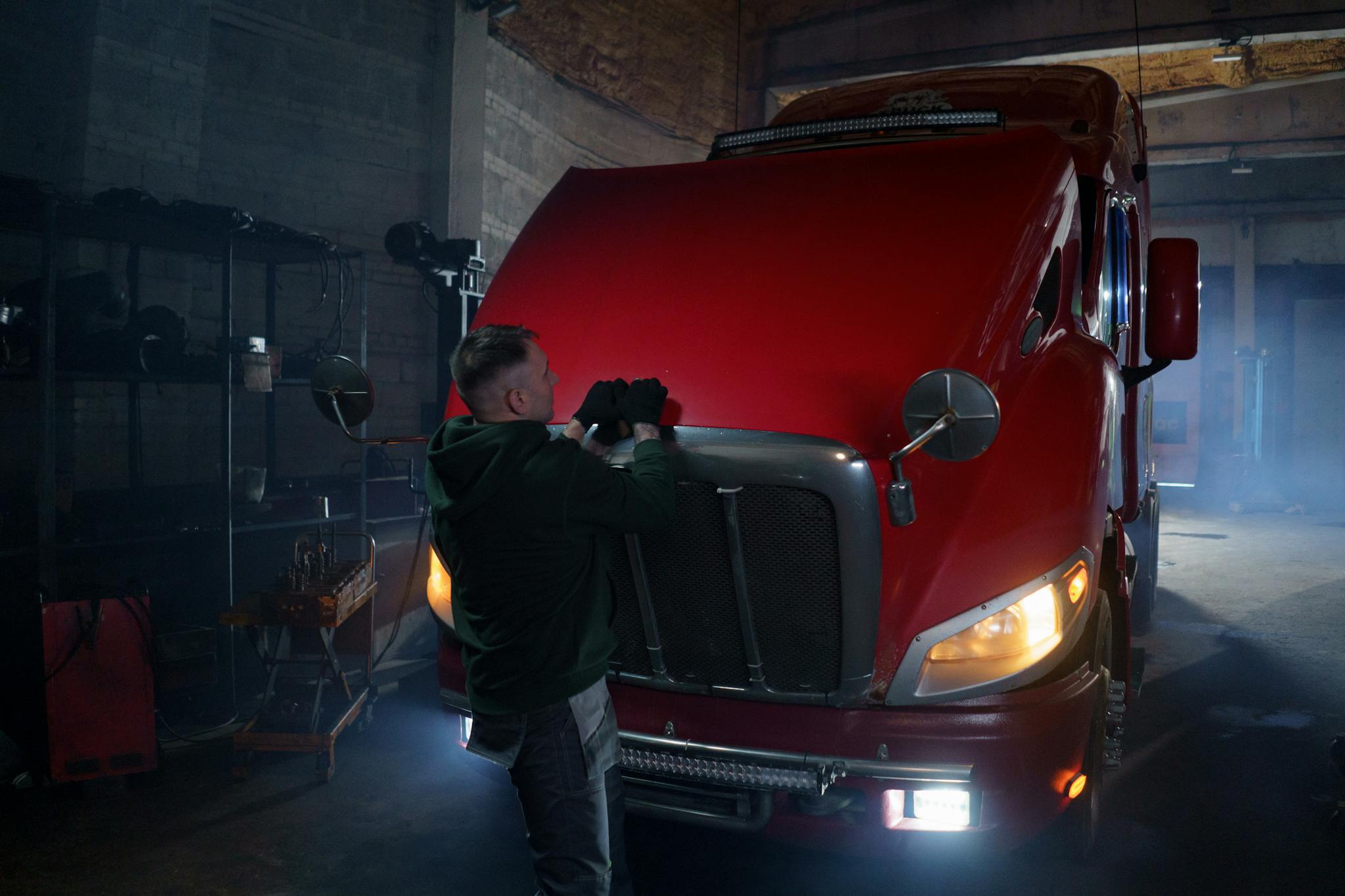 Mechanic wearing gloves working on red truck hood in dimly lit garage.